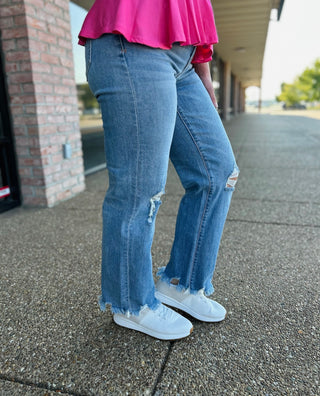 Woman wearing distressed wide leg jeans with frayed hem and white sneakers on concrete sidewalk.