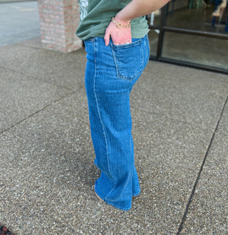 Person wearing medium wash wide leg palazzo jeans with green top and gold bracelet on sidewalk.