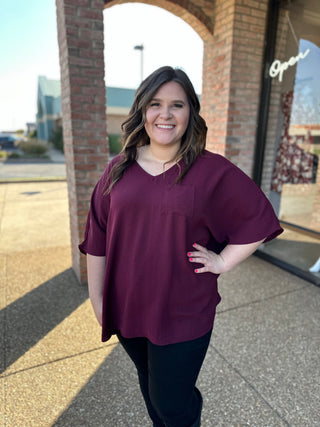 Woman wearing oversized burgundy v-neck blouse standing outdoors near brick archway storefront