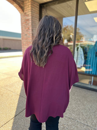Woman wearing oversized merlot purple v-neck blouse, shown from behind walking on sidewalk near storefront.