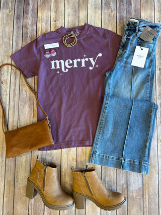 Purple "merry" graphic t-shirt styled with jeans, brown boots, purse and gold jewelry on wooden background.