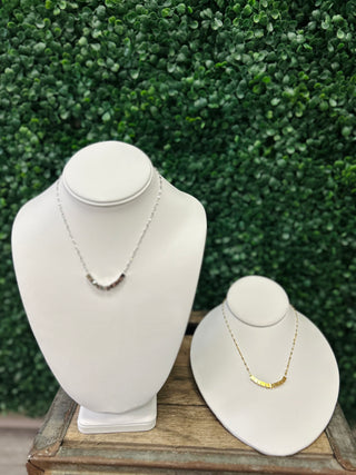 Two white jewelry display busts showcasing delicate necklaces on wooden surface with green foliage background.
