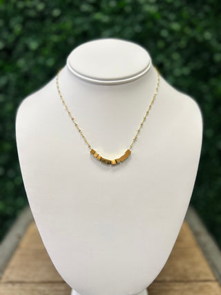 Gold cube pendant necklace displayed on white jewelry bust with green foliage background