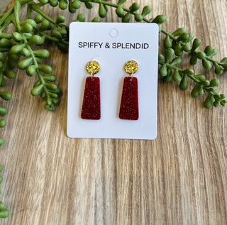 Sparkly red and gold dangle earrings on white card with green plants on wooden surface