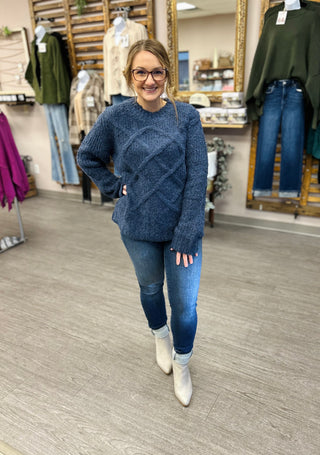 Woman wearing dark wash skinny jeans with blue sweater and white boots in boutique setting.