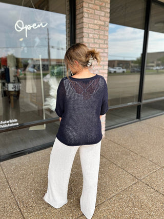 Woman seen from behind wearing a sheer crochet knit top paired with wide-leg pants outside boutique.