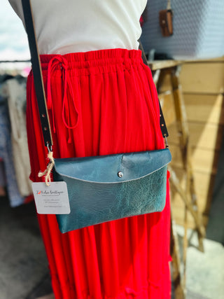 Person wearing red skirt modeling teal leather envelope crossbody bag with adjustable strap and brand tag.