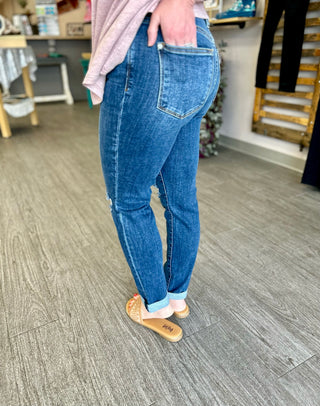 Person wearing blue denim jeans with distressed knee details and cuffed hem, standing on wood floor.
