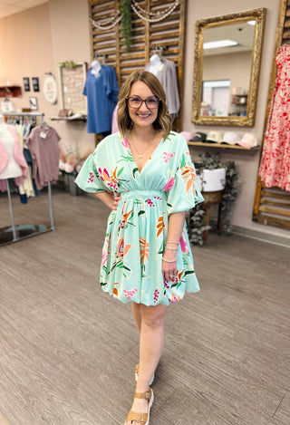 Woman wearing mint green floral dress with flutter sleeves in boutique clothing store setting.