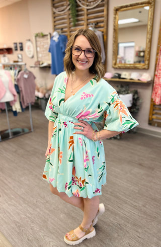 Woman wearing mint green floral dress with tropical print in boutique clothing store setting.