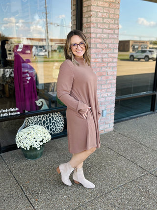 Woman wearing mocha mock neck sweater dress with boots, standing outside storefront with brick column and flowers.