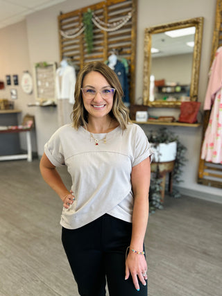 Woman wearing light gray asymmetrical top with short sleeves in modern retail boutique setting.