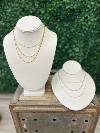 Two white jewelry display busts showcasing layered gold and silver chain necklaces on wooden crate outdoors.