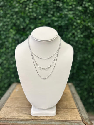 Three layered silver chain necklaces displayed on white jewelry bust against green foliage background.