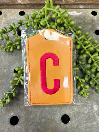 Orange embroidered monogram luggage tag with letter C in clear plastic packaging surrounded by green foliage