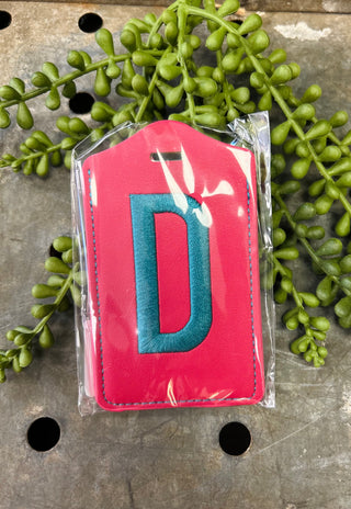 Red leather luggage tag with embroidered letter D in clear plastic packaging on decorative surface.