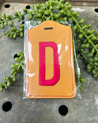 Orange leather luggage tag with embroidered red letter D in clear plastic packaging