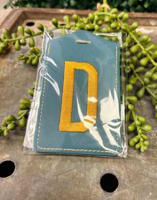 Embroidered monogram luggage tag in clear protective packaging surrounded by decorative greenery on wooden surface.