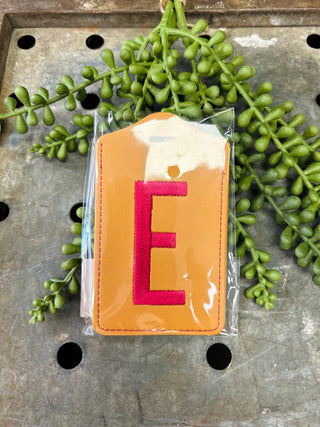 Orange leather luggage tag with red embroidered letter E in clear plastic packaging surrounded by green plants.