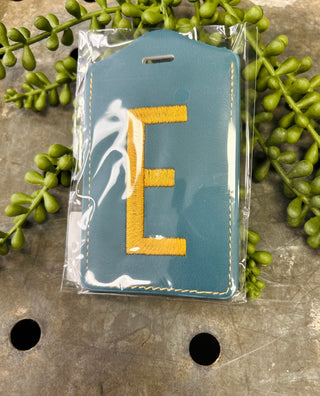 Ganz monogram luggage tag with embroidered letter in clear packaging surrounded by green succulent plants.