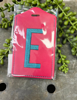 Pink luggage tag with embroidered blue letter E in clear protective packaging surrounded by green foliage.