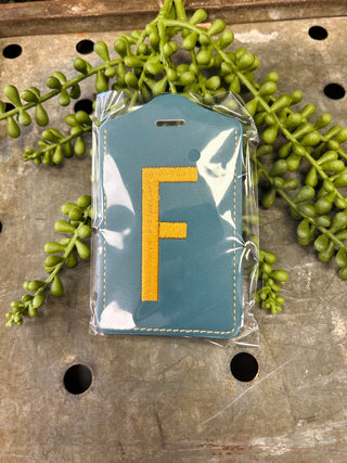 Blue leather luggage tag with embroidered letter F in clear plastic packaging, surrounded by green foliage.