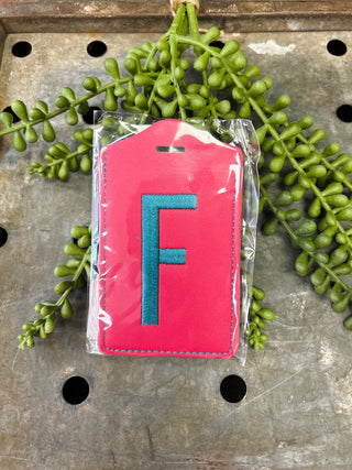 Red leather luggage tag with embroidered letter F in clear plastic packaging on decorative surface.