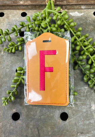 Orange leather luggage tag with embroidered red letter F, packaged in clear plastic bag.