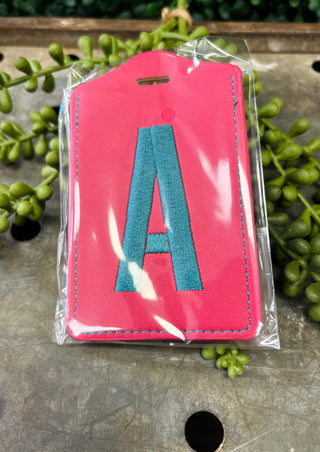 Pink leather luggage tag with embroidered letter A in clear protective packaging on wooden surface.
