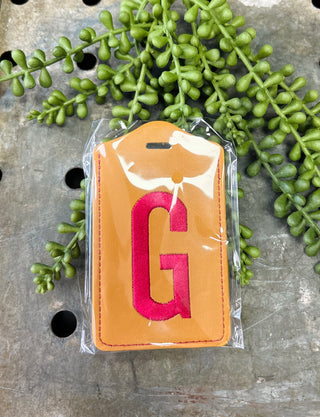 Orange leather luggage tag with red monogram letter G in clear plastic packaging surrounded by green foliage.