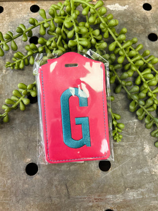 Red leather monogram luggage tag with letter G displayed on metal surface with green plants