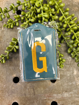 Embroidered monogram luggage tag in clear protective packaging surrounded by decorative greenery