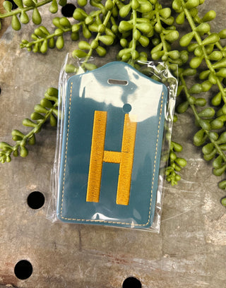 Monogram luggage tag with embroidered letter H in clear packaging surrounded by green foliage