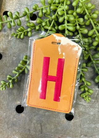 Orange leather luggage tag with red monogram letter H in clear protective packaging