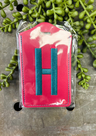 Pink leather luggage tag with embroidered letter H in clear protective packaging surrounded by greenery.