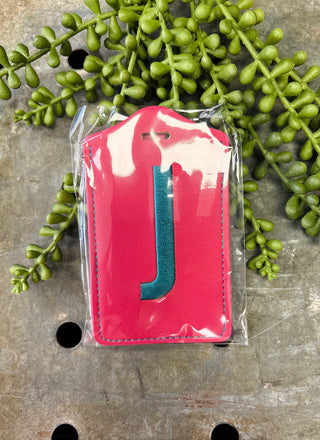 Red leather luggage tag with monogram letter J in clear plastic packaging surrounded by green foliage.