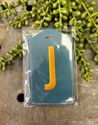 Ganz monogram luggage tag with embroidered letter in clear packaging surrounded by greenery