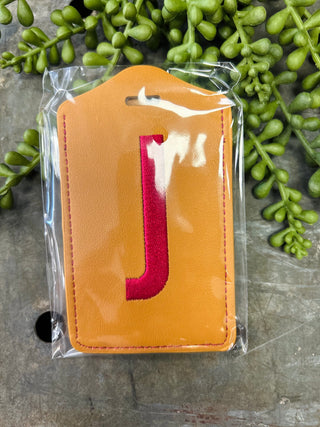 Orange leather luggage tag with red monogram letter in clear plastic packaging on stone surface.