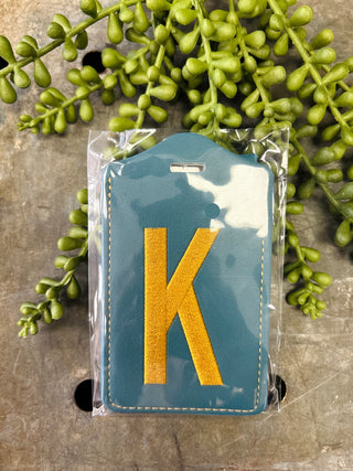 Blue leather luggage tag with embroidered letter K surrounded by green succulent plants on wooden surface.