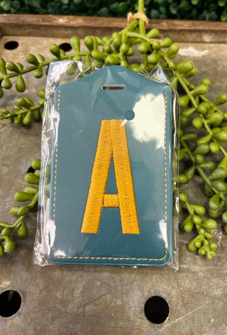 Monogram luggage tag with embroidered letter on blue background, packaged in clear plastic wrapper.