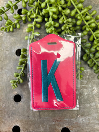 Monogram luggage tag with letter K in clear protective packaging surrounded by green foliage