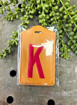 Orange leather luggage tag with red monogram letter K in clear protective packaging surrounded by green foliage.