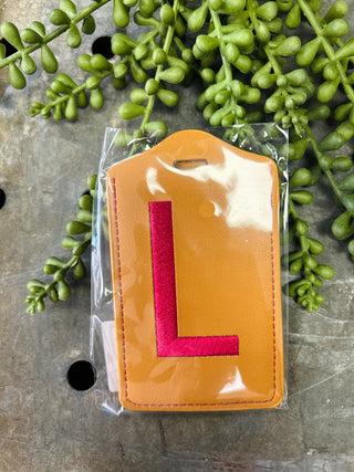 Orange leather luggage tag with red monogram letter in clear plastic packaging surrounded by green plants.