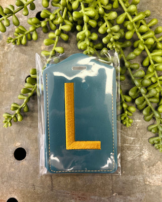 Monogram luggage tag with embroidered letter in clear protective packaging surrounded by green foliage