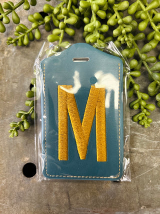 Monogram luggage tag with letter M in clear protective packaging surrounded by green succulent plants