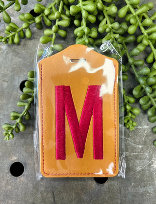 Orange leather luggage tag with red monogram letter M in clear plastic packaging surrounded by green plants.