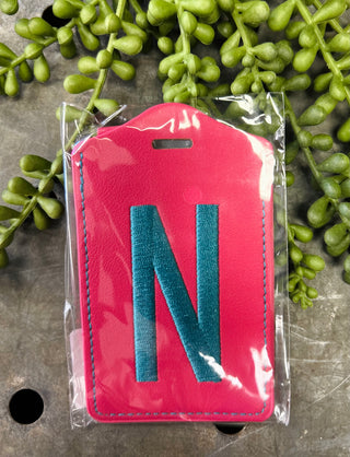 Pink leather luggage tag with monogram letter N in clear plastic packaging surrounded by green foliage.