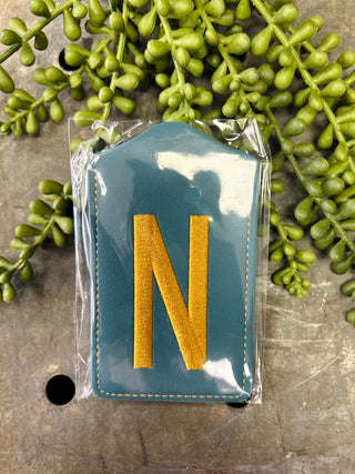 Blue leather luggage tag with embroidered letter N surrounded by green foliage on stone surface