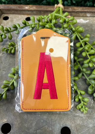 Leather luggage tag with embroidered monogram letter in clear protective packaging on wooden surface