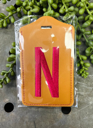 Leather luggage tag with embroidered letter N in clear plastic packaging surrounded by greenery